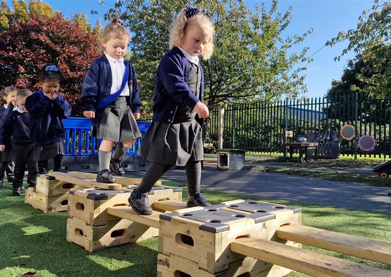 Wooden planks and blocks are connected in a straight line to create a simple obstacle course. 7 children are waiting in a single-file line as each child walks carefully across the obstacle course.